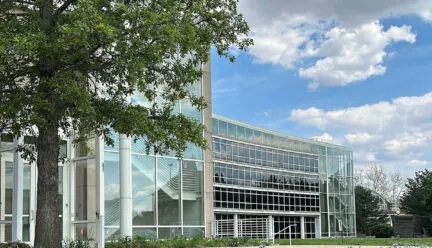 building with green trees, blue sky with white clouds, and garden with purple flowers surrounding it