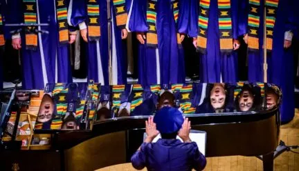 chorus wearing blue robes singing and reflected in black piano