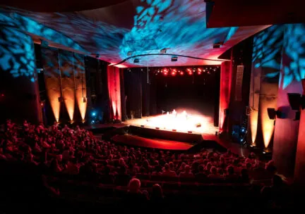 interior of a theatre proscenium with orange and blue lights on the ceiling and onstage and many people seated in the auditorium