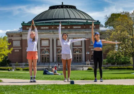 three students on the central green grassy quad hold up their arms to make "I-L-L"