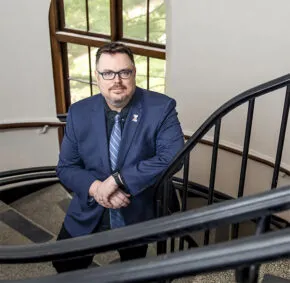 man with brown hair, facial hair, glasses, in a dark gray suit with lavender shit