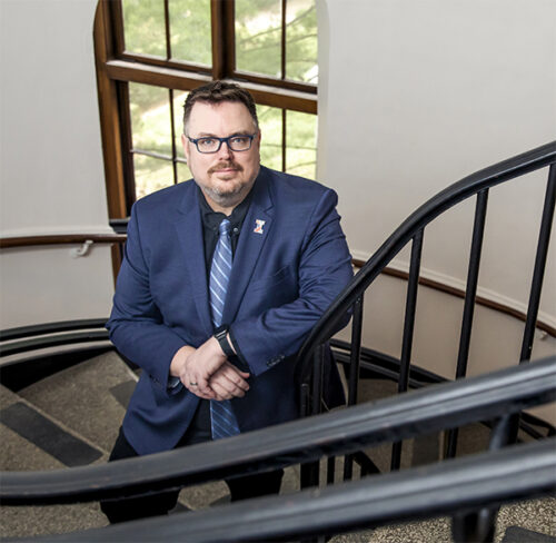 man with brown hair, facial hair, glasses, in a dark gray suit with lavender shit