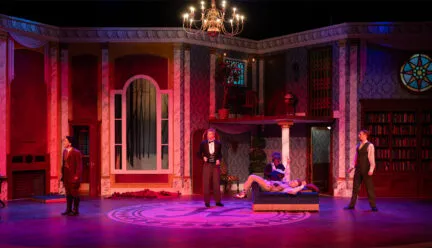 stage set that looks like a luxurious early 20th century home drenched in pink and purple lighting with actors onstage