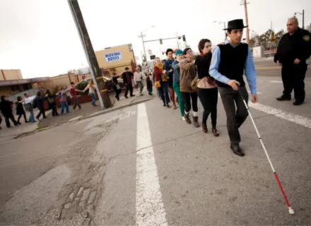 a person uses a white cane on a road while a line of people hold on one by one with their eyes closed