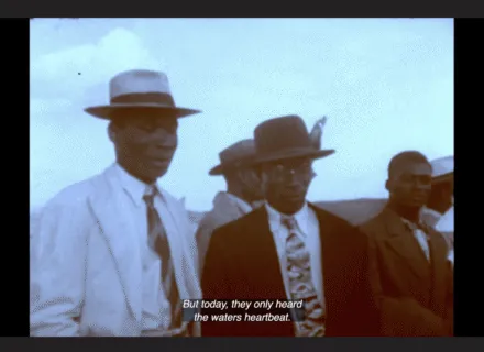 A group of Black men are standing next to each other. They are dressed in suits, ties and hats. Above them is a wide expanse of sky. The colors in this still image from Handsworth Songs are very muted, almost black and white. The captions at the bottom of the screen read "But today, they only heard the waters heartbeat."
