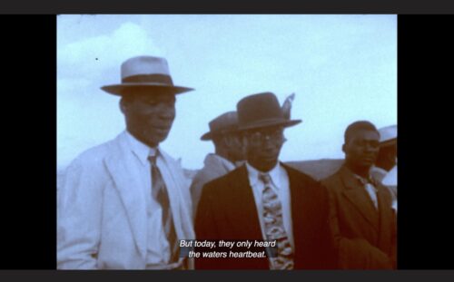 A group of Black men are standing next to each other. They are dressed in suits, ties and hats. Above them is a wide expanse of sky. The colors in this still image from Handsworth Songs are very muted, almost black and white. The captions at the bottom of the screen read "But today, they only heard the waters heartbeat."