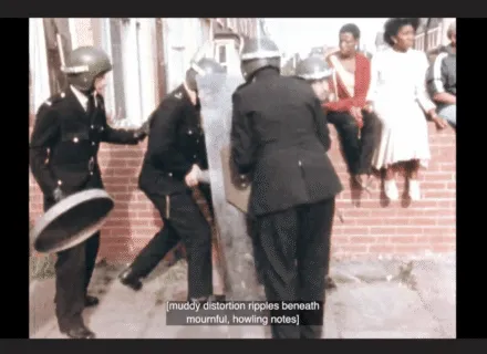A still image from the film Handsworth Songs. A group of three white policemen equipped with shields and helmets surround someone. Behind them on a brick wall three Black people are sitting. The captions at the bottom of the image read “[muddy distortion ripples beneath mournful, howling notes]”.