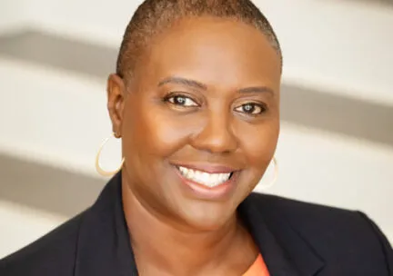 woman smiling at the camera, close shave head, large earings, black blazer, orange shirt