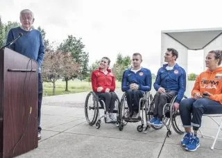 speaker at lectern, three people sitting in wheelchairs, and one person sitting in a chair