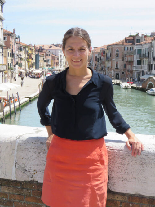 Elisa Silva in Italy, standing a bridge in front of houses, river inbetween