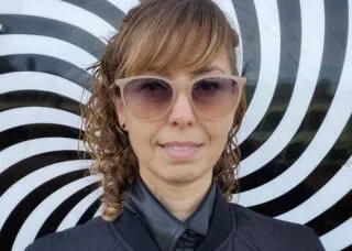 blond woman with sunglasses smiling at camera with straight and curly hair blue zipped up rain jacket and blakc and white rotating background