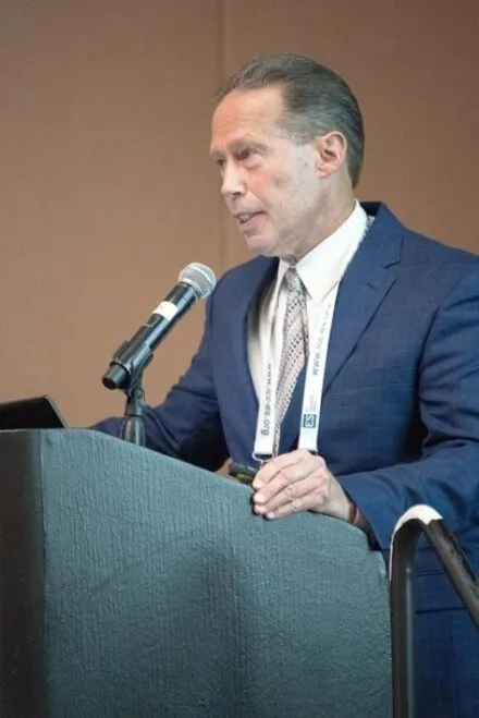man speaking a lecturn, hair slicked back, white complexion, blue blazer, white button up shirt