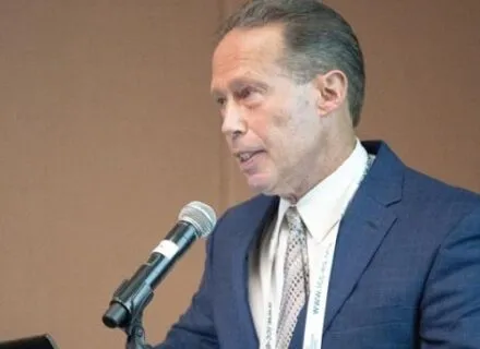 man speaking a lecturn, hair slicked back, white complexion, blue blazer, white button up shirt