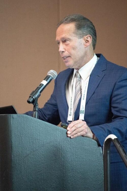man speaking a lecturn, hair slicked back, white complexion, blue blazer, white button up shirt