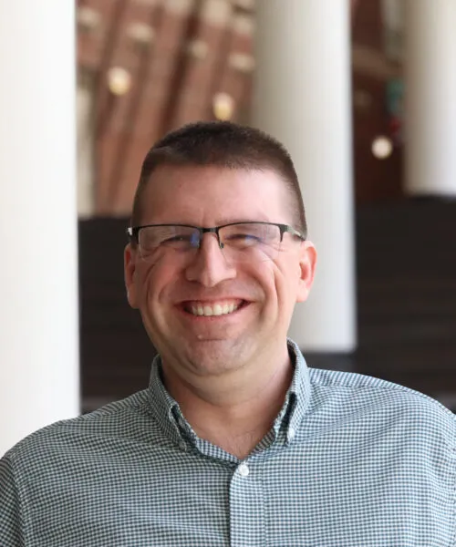 man smiling at camera, green shirt black framed glasses black hair short crop
