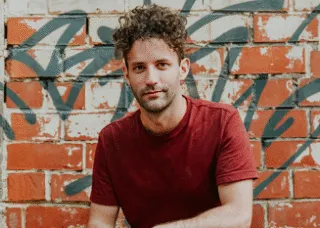 man looking at camera with graffiti behind him squatting with jeans and red shirt