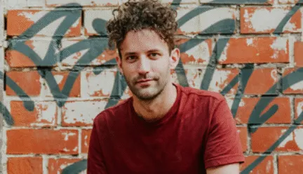 man looking at camera with graffiti behind him squatting with jeans and red shirt