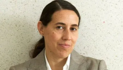 woman with shoulder length hair in ponytail wearing beige suit with white button up and beige vest with a knowing smile on her face looking at camera