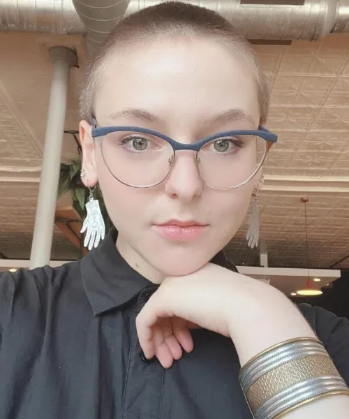 woman looking at camera, bald, blue framed glasses black shirt hand under chin