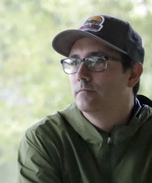 Portrait of Ryan Griffis in an outdoor setting, wearing a baseball hat and green rain jacket.