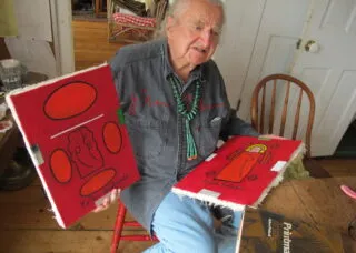 Peter Bodnar sitting on chair holding two of his paintings in bold red
