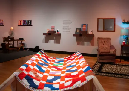 a museum installation shows a colorful quilt in the foreground, with other furniture pieces in the background