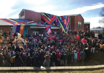 hundreds of children gather around their school as it is covered with colored fabric