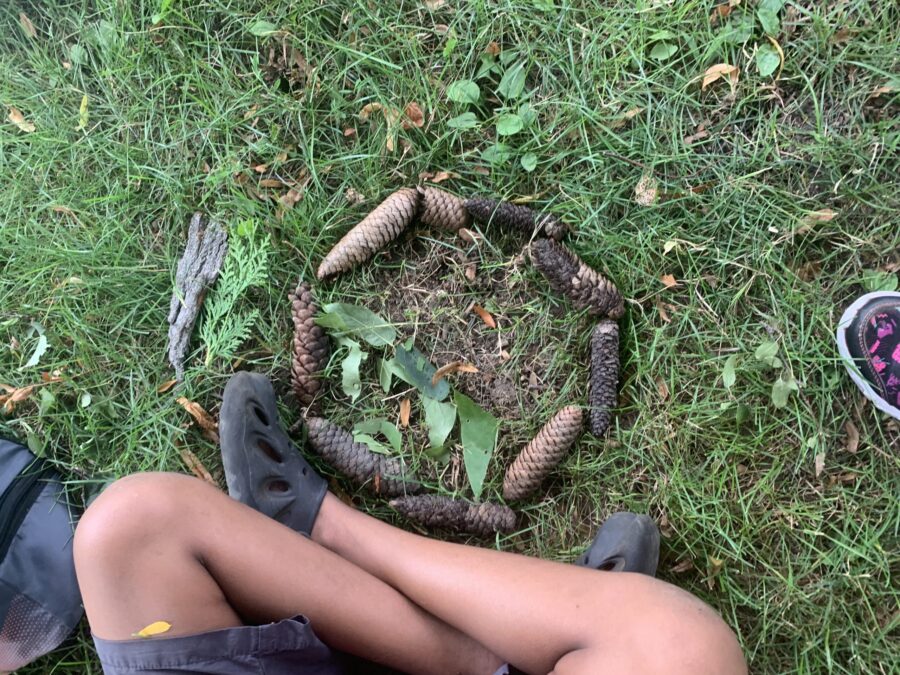 using pinecones, a child creates a circle sculpture on the grass. their legs are in the picture. 