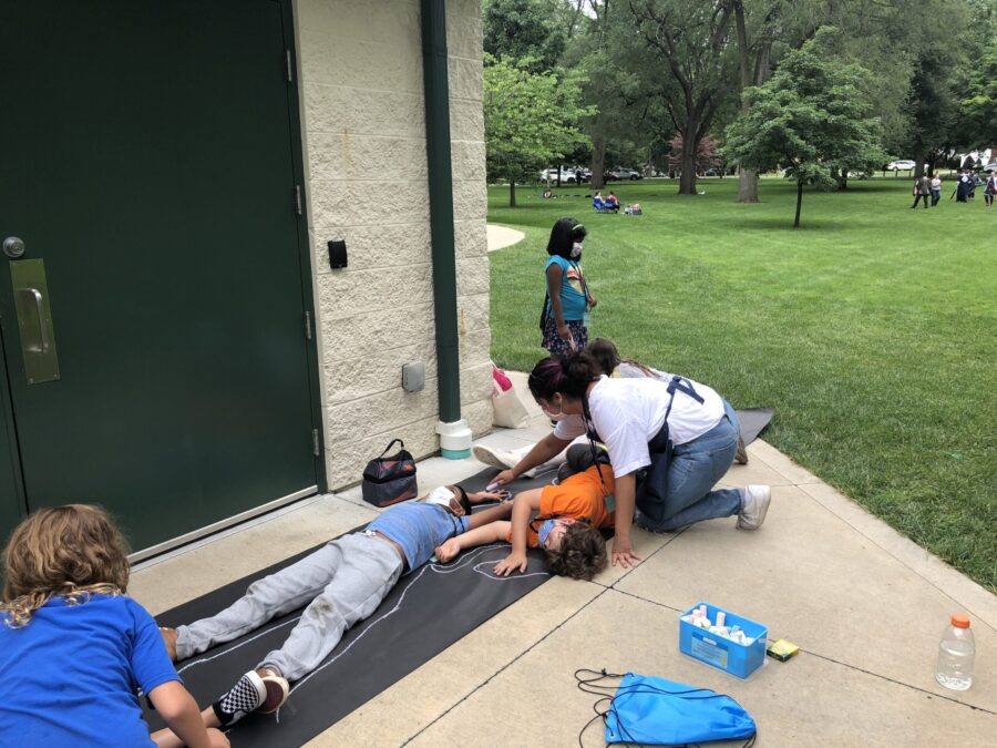 children trace each other's bodies with chalk on a large black piece of paper, outdoors