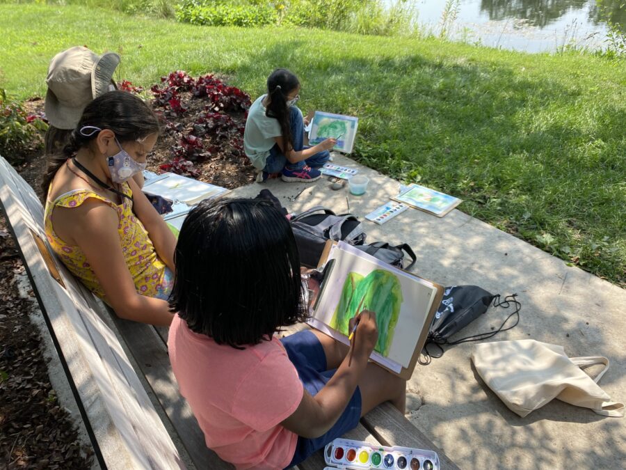 a group of young students paints with watercolors on the bank of a pond, shadows are cast from the trees over the kids