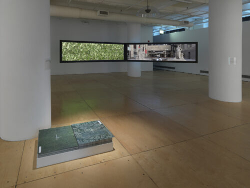 A 10 foot wide by 2 feet tall video projection showing landscape images is suspended about five feet off of a wood gallery floor. In the foreground, two stacks of maps that are eleven by seventeen inches each sits on the floor next to one another.