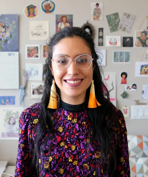 brown skinned femme, wearing big white glasses, long yellow earrings and a colorful shirt with flowers