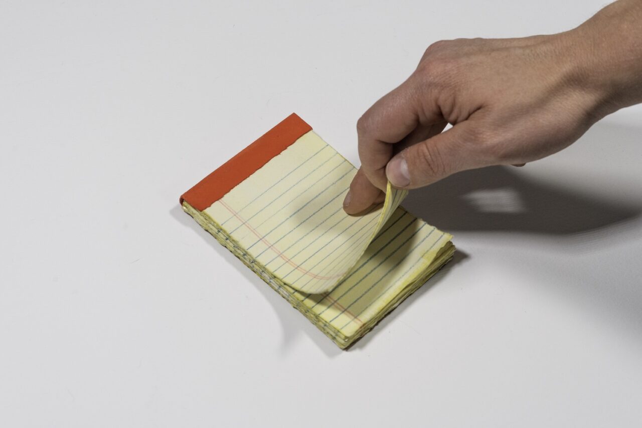 A hand is flipping pages on a 5 by 3 and one half inch ruled yellow notepad, against a white background.