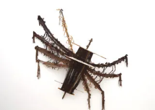 [ID: A multimedia textile where the artist has handwoven brown threads into the shape of a large spider. The artist has incorporated elements of the loom to add to the body and structure of the spider.]