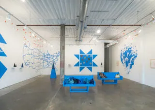 [ID: A gallery with white walls featuring a design of triangles, geometric shapes, an outline of the US, and an organic shape resembling an enormous puddle; all are painted in the same medium-blue color. Along the walls are shelves with cone-shaped sculptures and small orange-and-white flags. In the center of the space are two medium-blue benches covered with indecipherable objects.]