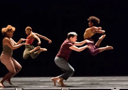 Two dancers arch their backs in a jump behind two dancers gesturing with their hands in front of their body