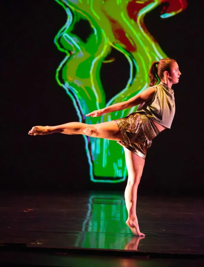 A lone dancer reaches their right arm forward in front of a green scrim