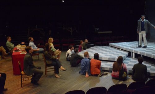 Young person standing center stage addressing audience seated mostly on the floor.