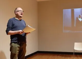 Andrei Kureichik wearing a blue sweater and glasses, stands holding a script, read aloud the name of the next speaker, while a projected image of a person shines behind him.