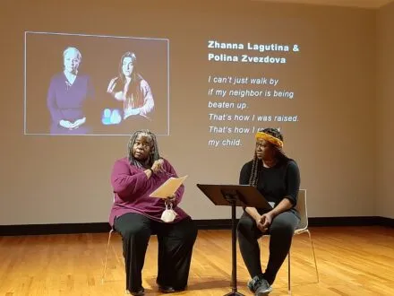 Two women dressed in dark blacks and purples sit in chairs reading from scripts while a projection of two other women and their names is shown behind them