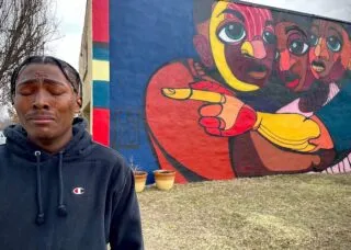 image of a person in a blue sweater crying in front of a mural depicting 3 people hugging