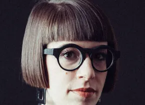 a person wearing a black jacket and thick dark-rimmed glasses looks out