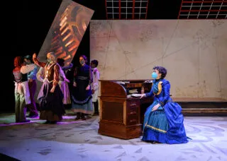 one actor wearing a blue dress sits at a wooden desk. other actors stand on the other end of the stage in a circle