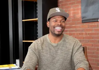 tyrone phillips smiles while sitting in a chair with a brick wall background and black bookshelves