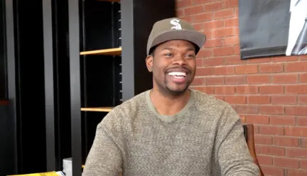 tyrone phillips smiles while sitting in a chair with a brick wall background and black bookshelves