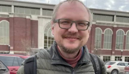 andrei, weaing glasses and a grey zip-up coat, holds a black and grey book and smiles