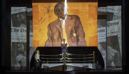 stage with actors around a boxing ring and a large painted backdrop featuring jack johnson with newspaper text across