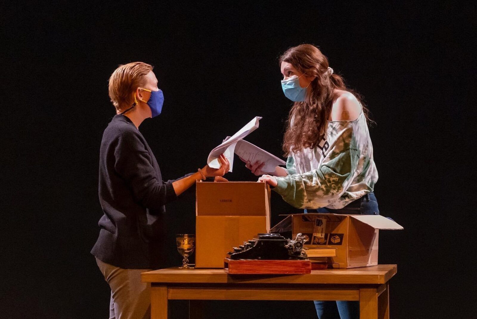 two actors wearing masks with a table and boxes around them