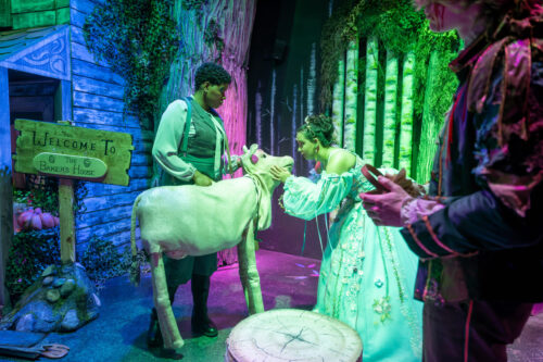 actors onstage, one holds a cow puppet and one pets it