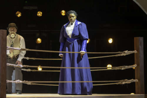 person onstage in blue dress walking in wrestling rink from the early 1900s